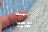 May include: A small, gold-colored arrow charm with a detailed feather design on one end and a diamond shape on the other. The charm has small loops on each end. Text on the image reads "14K Gold Filled Made in USA."