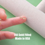 May include: A tiny gold-colored charm shaped like a pair of sunglasses with small loops on each side for attachment. The charm is 14K gold filled and made in USA. The metal piece is shown resting on a finger against a textured fabric background.